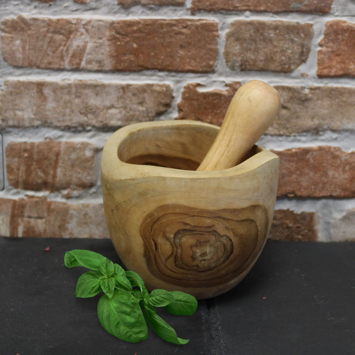 Mortero y maja de madera de teca con veta natural, estilo rústico y artesanal, junto a unas hojas de albahaca fresca.