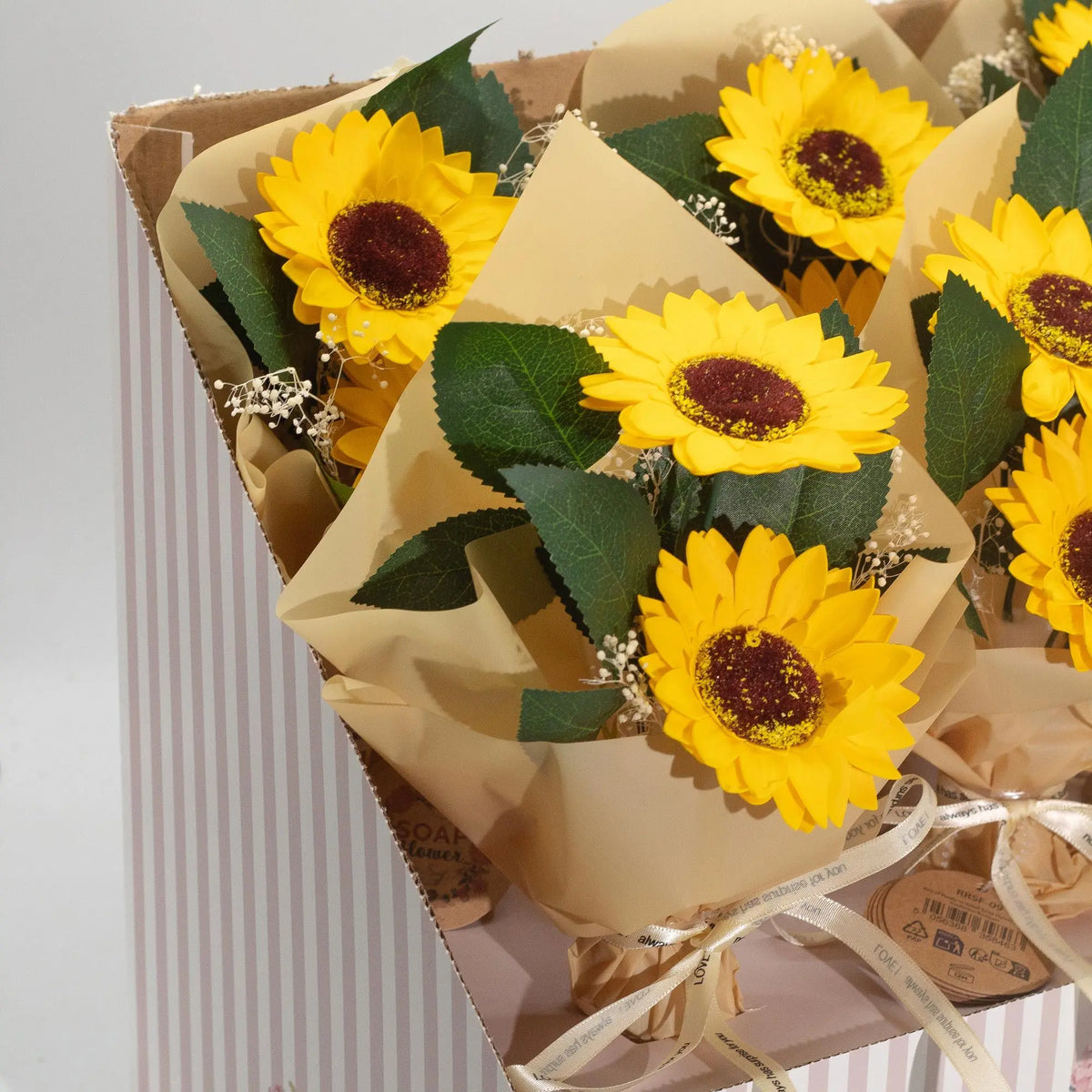 Ramo de dos girasoles hechos de jabón, con pétalos amarillos, hojas verdes, envueltos en papel kraft y cinta beige.