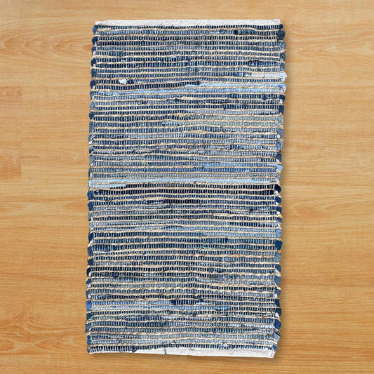 Alfombra rectangular tejida en yute y algodón reciclado azul y crudo, sobre un suelo de madera claro.