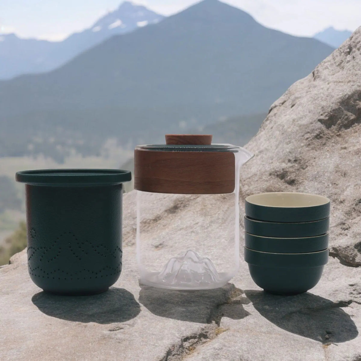 Set de té portátil: tetera de vidrio con tapa de madera, infusor y 4 tazas verde oscuro sobre roca, fondo de montaña.