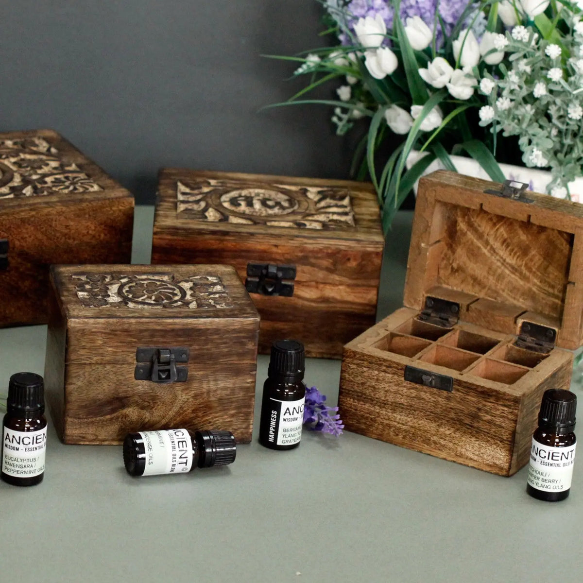 Cajas de madera de mango tallada con compartimentos para aceites esenciales y cierre metálico, sobre fondo neutro.