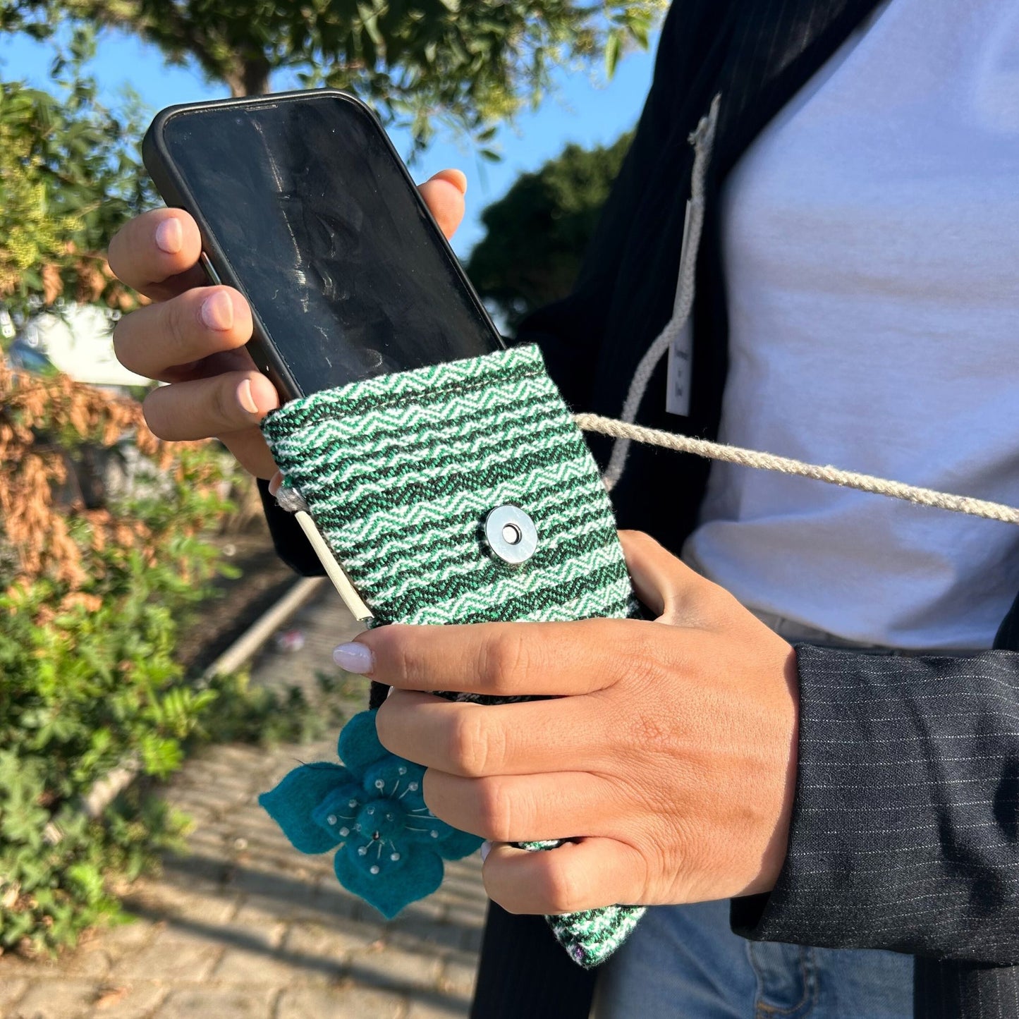 Bolso porta teléfono de tejido verde a rayas con botón metálico, cordón y colgante de flor azul.