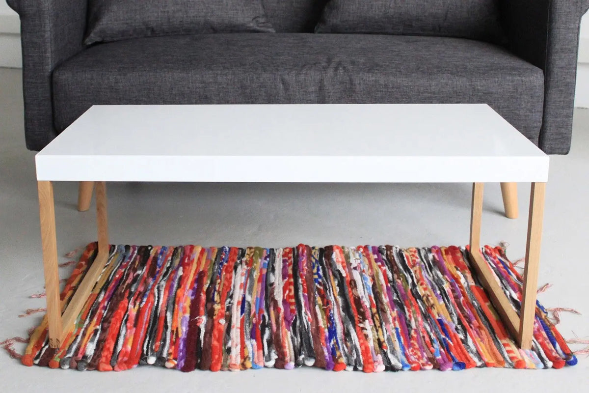 Alfombra de trapo artesanal, rectangular y multicolor, bajo una mesa de centro blanca con patas de madera clara.
