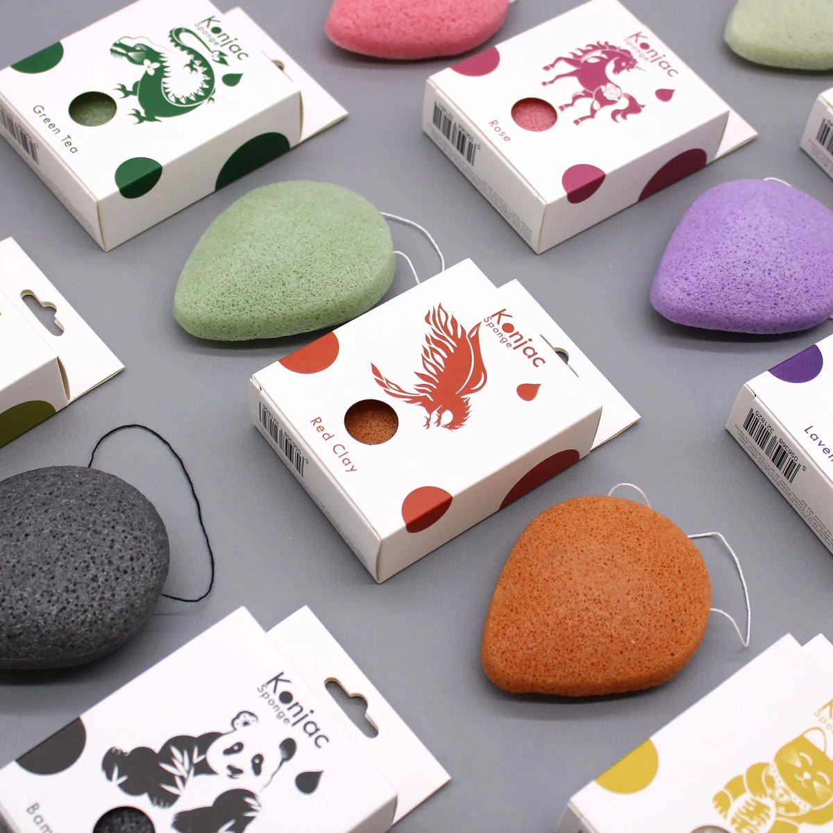 Esponjas faciales Konjac de colores y formas variadas con sus cajas de empaque sobre fondo gris.