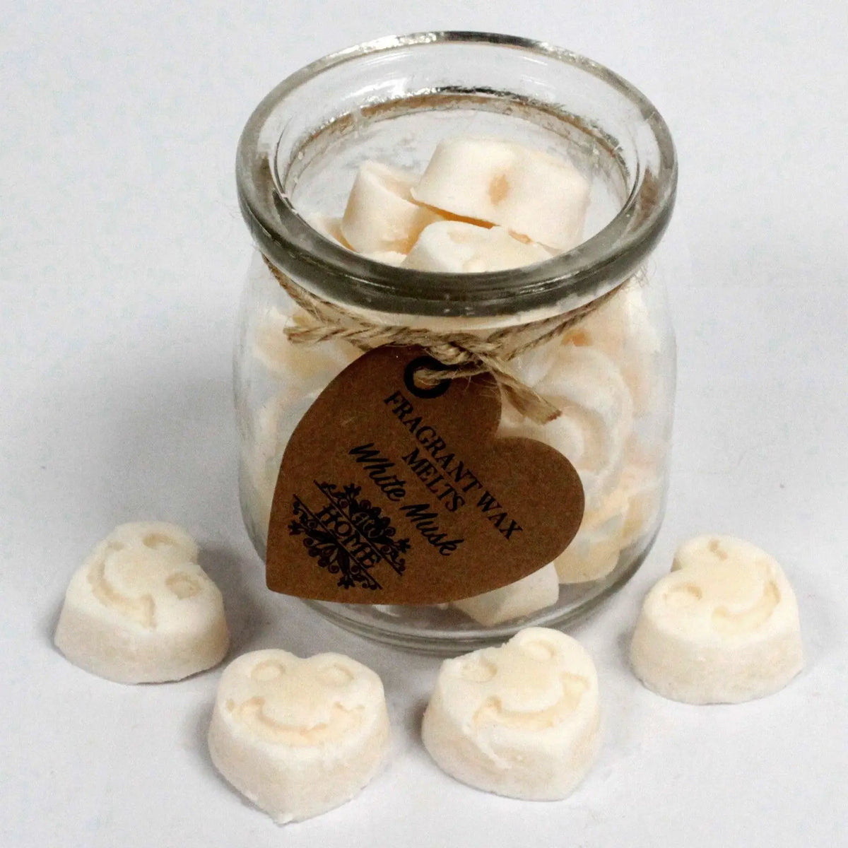 Frasco de cristal con cera de soja en forma de carita feliz y etiqueta en forma de corazón sobre fondo blanco.
