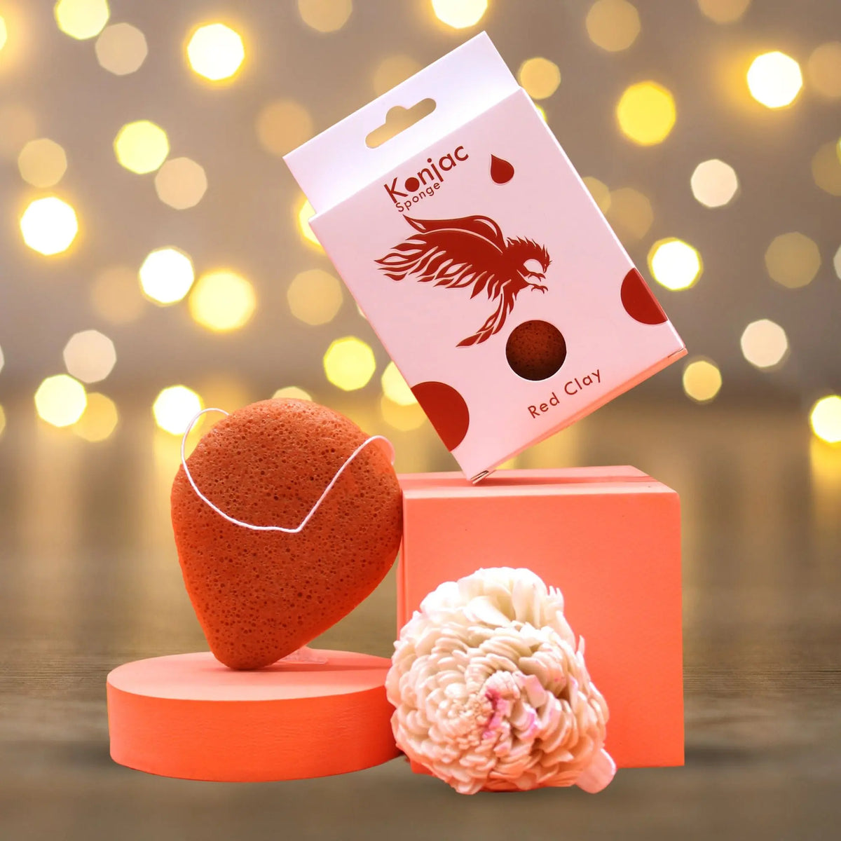Esponja Konjac de arcilla roja sobre bases naranjas con caja y flor decorativa frente a luces cálidas desenfocadas.