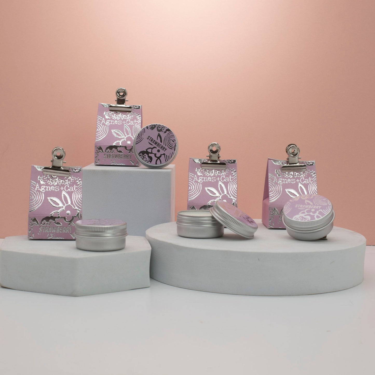 Bálsamos labiales Agnes+Cat de fresa en latas metálicas y bolsas de papel rosa con detalles plateados sobre pedestales.