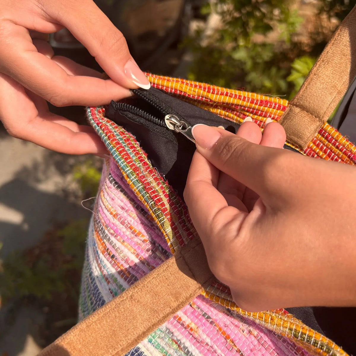 Manos abriendo bolso grande de tejido reciclado multicolor en tonos rosa, amarillo y naranja con cremallera negra.