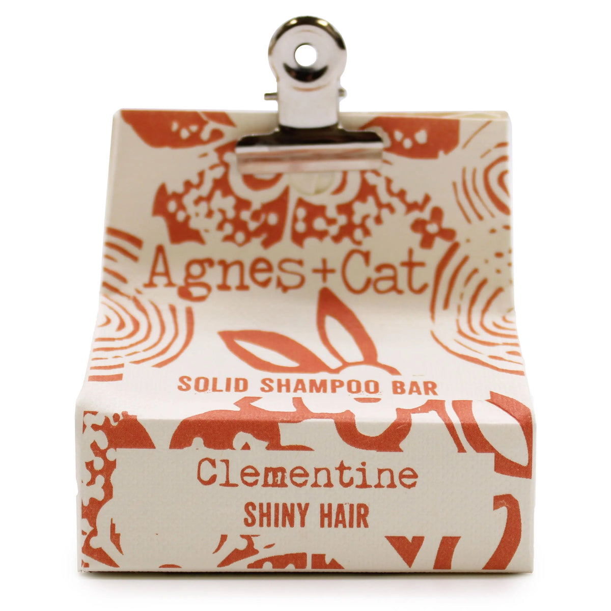 Champú sólido Clementina Agnes+Cat en caja de papel crema con estampado naranja, sujeto por una pinza metálica superior.