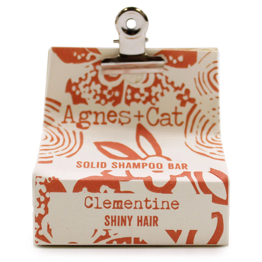Champú sólido Clementina Agnes+Cat en caja de papel crema con estampado naranja, sujeto por una pinza metálica superior.
