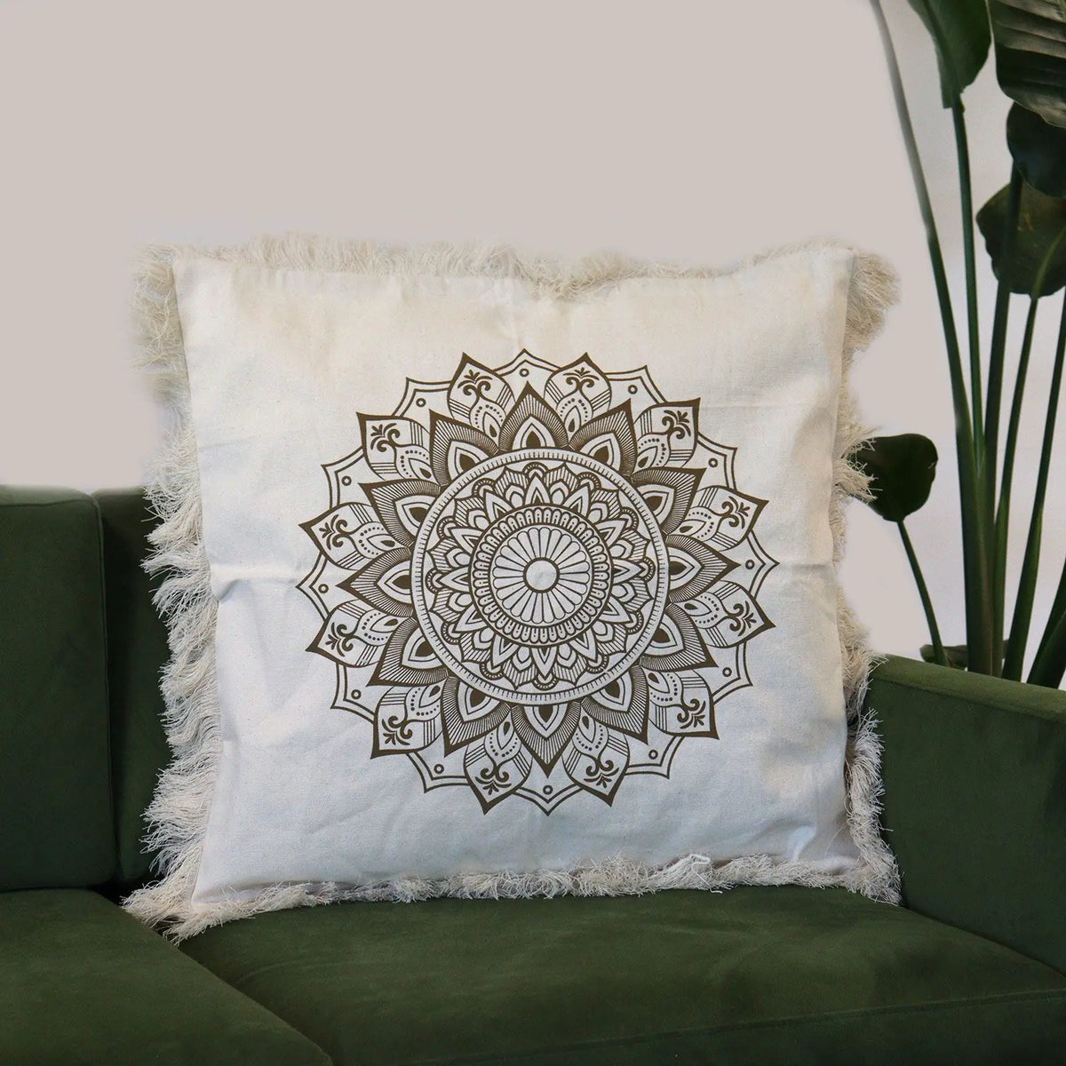 Funda de cojín beige cuadrada con diseño de mandala en bronce y bordes decorados con flecos, sobre un sofá verde.