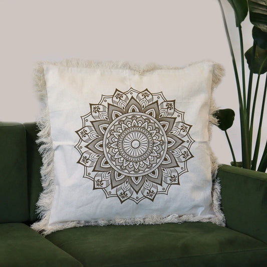 Funda de cojín beige cuadrada con diseño de mandala en bronce y bordes decorados con flecos, sobre un sofá verde.