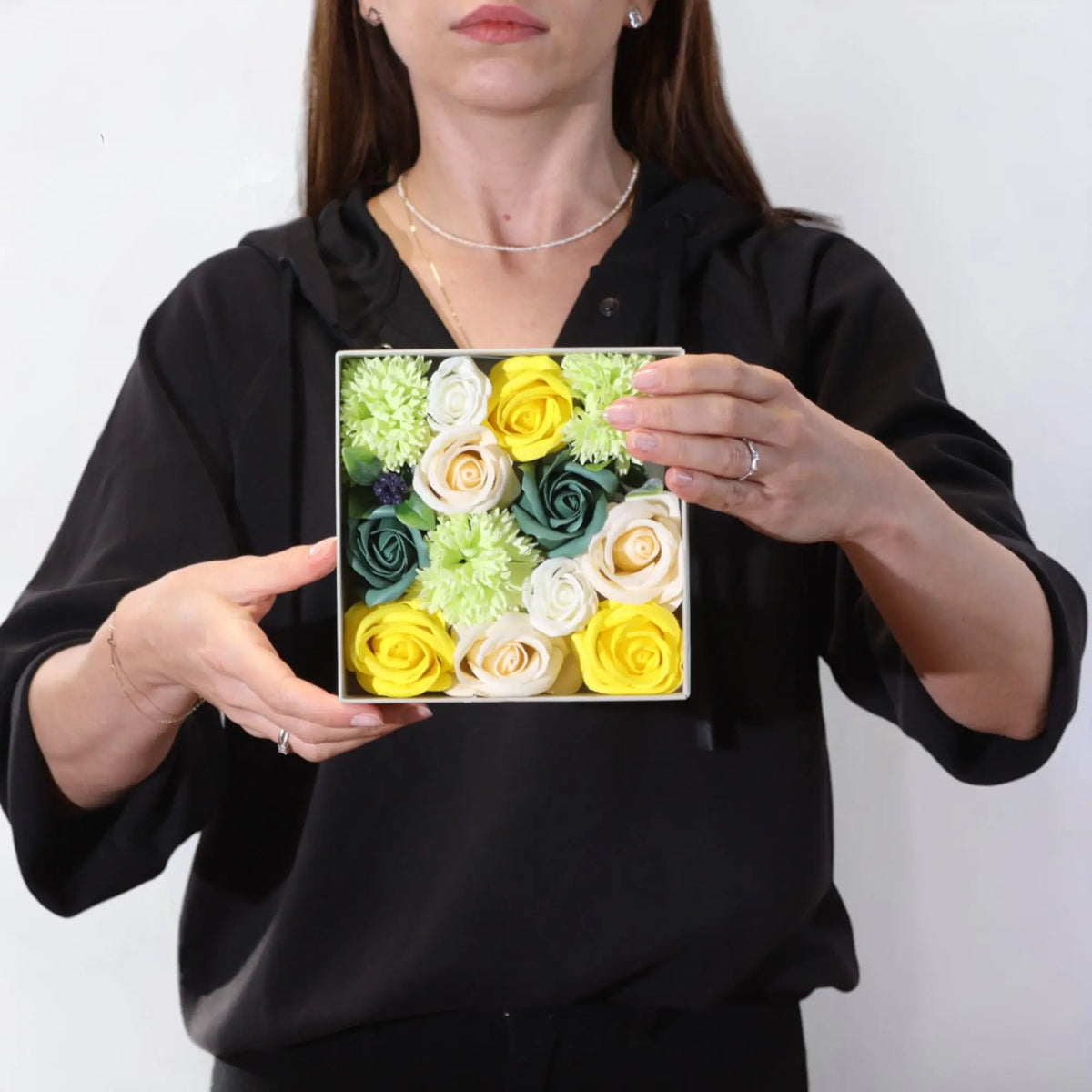 Caja cuadrada con flores de jabón amarillas, blancas y verdes, sostenida por una persona con ropa negra.