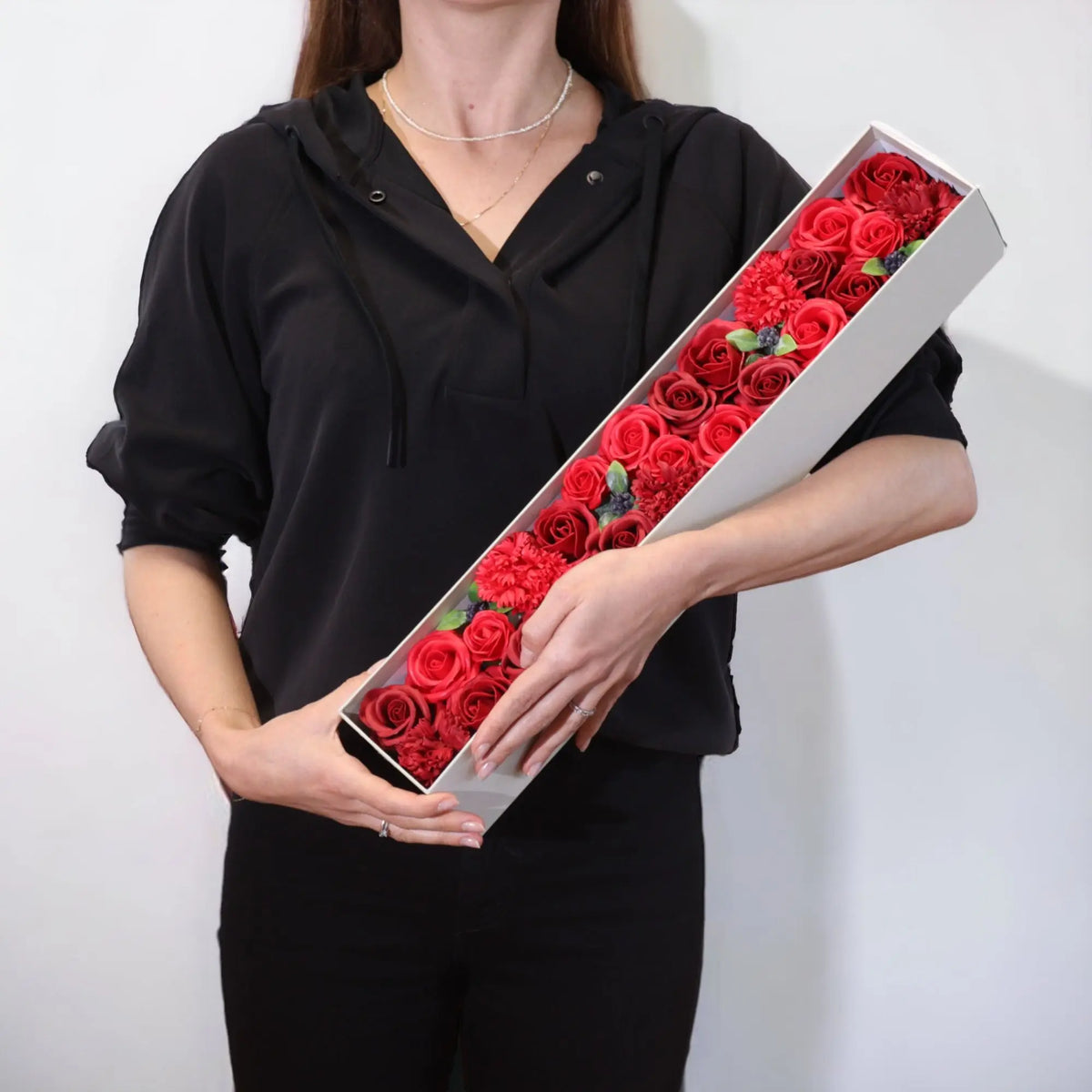 Persona sosteniendo una caja rectangular alargada llena de rosas y flores de jabón rojas.