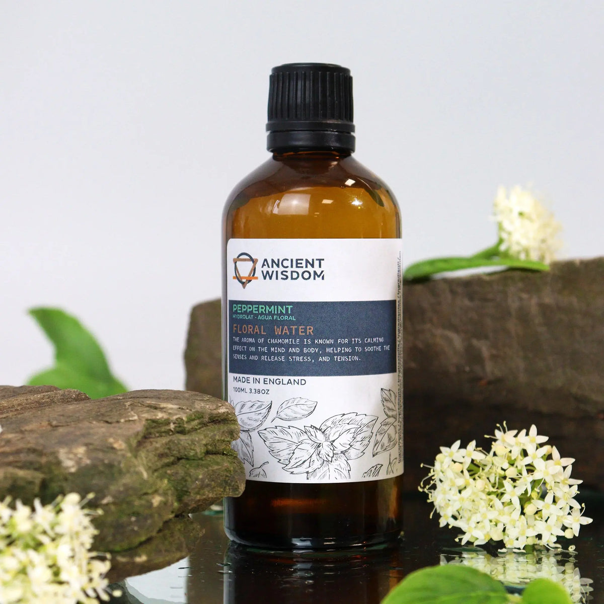 Frasco de vidrio ámbar de 100ml con hidrolato de menta sobre base de piedra, rodeado de flores blancas.