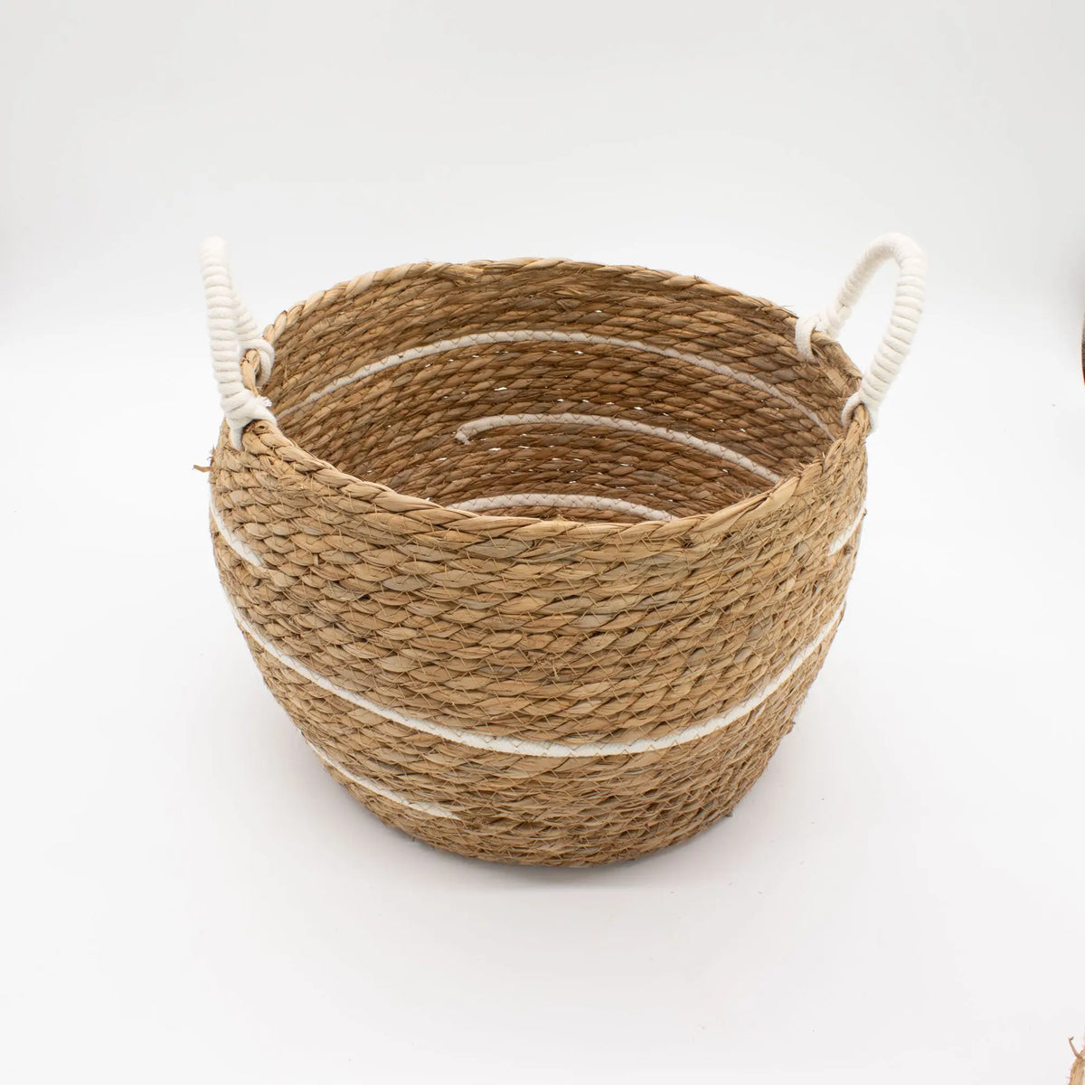 Cesta redonda de pasto marino trenzado con rayas de algodón blanco y asas laterales de cuerda, sobre fondo blanco.