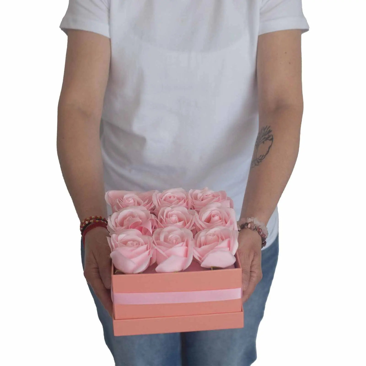 Persona sosteniendo una caja cuadrada color rosa con 9 rosas de jabón rosadas, adornada con una cinta satinada.
