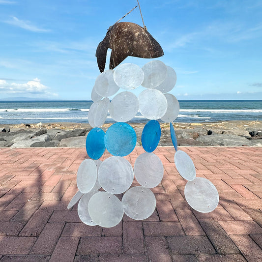 Campanilla de viento con base de coco y discos de nácar traslúcidos blancos y azules, frente a una playa.