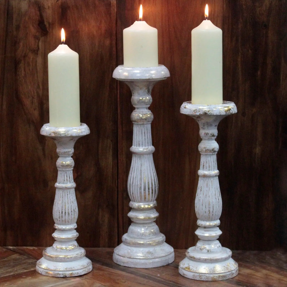 Tres candelabros de madera vintage en blanco con detalles dorados, cada uno con una vela blanca encendida.