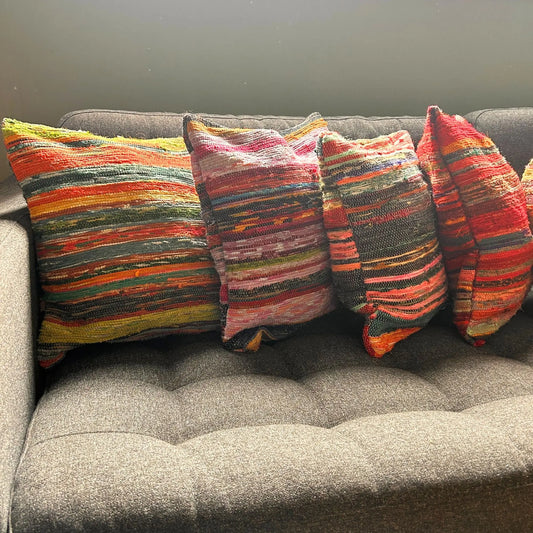 Cuatro fundas para cojín de 40x40cm hechas de tela reciclada a rayas multicolores, sobre un sofá gris.
