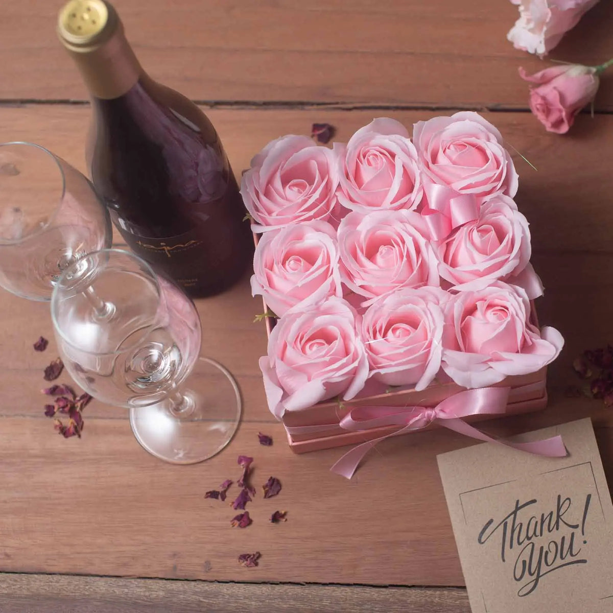 Caja cuadrada rosa con 9 rosas de jabón, junto a una botella de vino, dos copas y una tarjeta de agradecimiento.