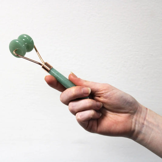 Mano sosteniendo un rodillo facial de jade verde con doble esfera y mango de metal en tono oro rosa. Fondo blanco.