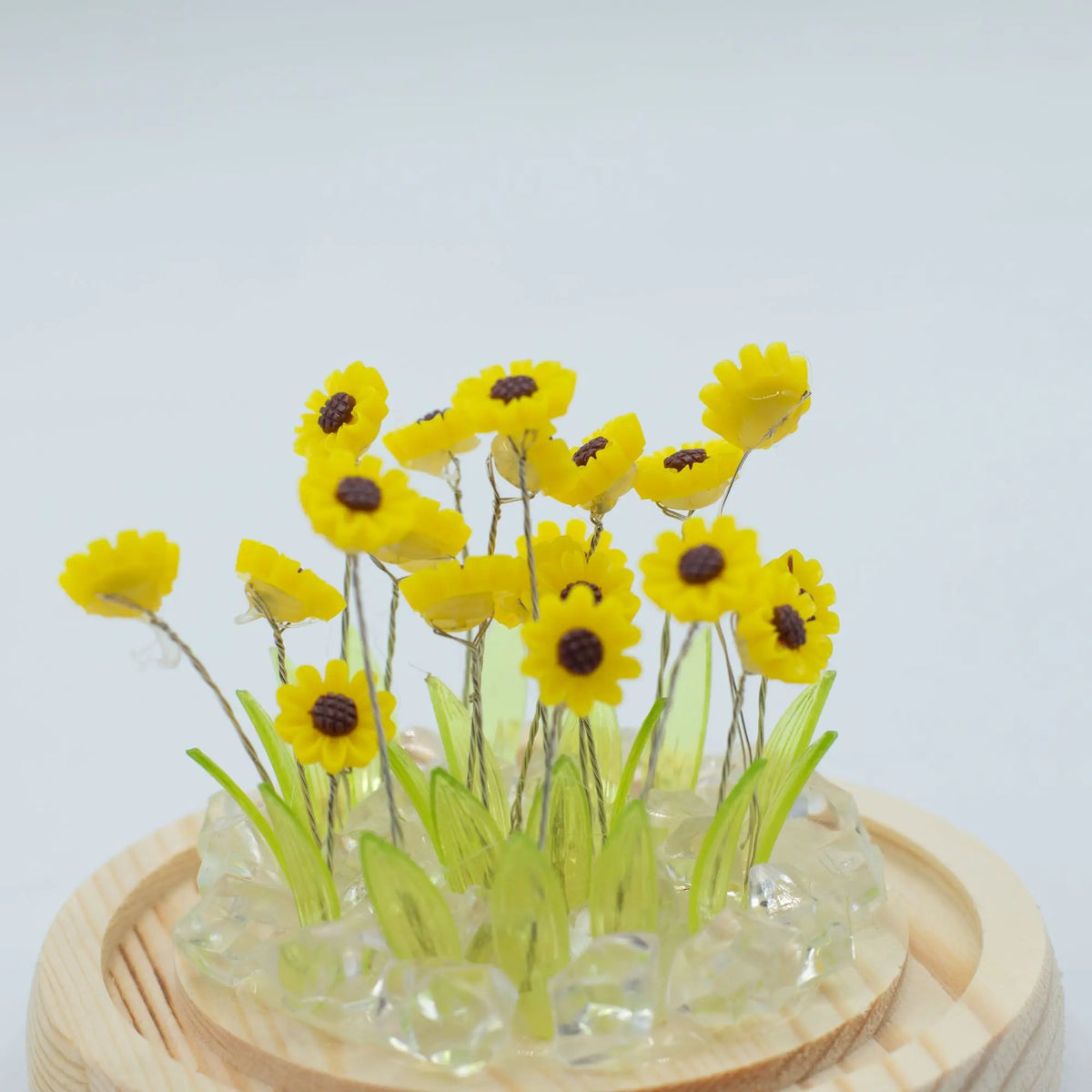 Domo Floral Glow: 20 mini girasoles amarillos de plástico, hojas verdes y cristales sobre base circular de madera.