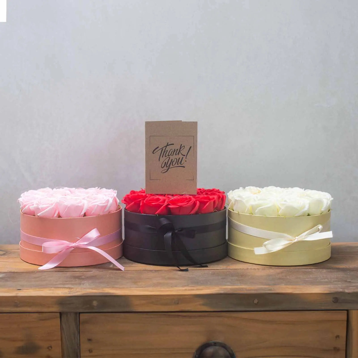 Tres cajas redondas con rosas de jabón (rosas, rojas y blancas) con lazos y una tarjeta de agradecimiento sobre madera.