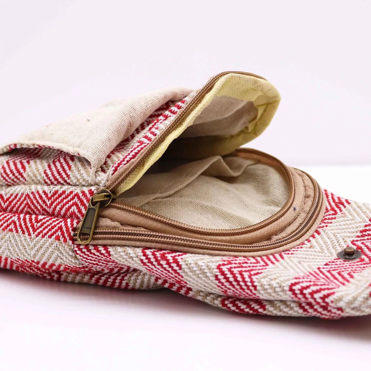 Bolso bandolera de algodón con tejido de zigzag en beige y rojo, con cierre de cremallera y solapa sobre fondo blanco.