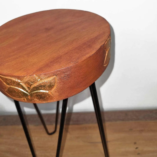 Soporte para plantas de madera redonda color terracota con flores talladas y patas de metal negro estilo horquilla.