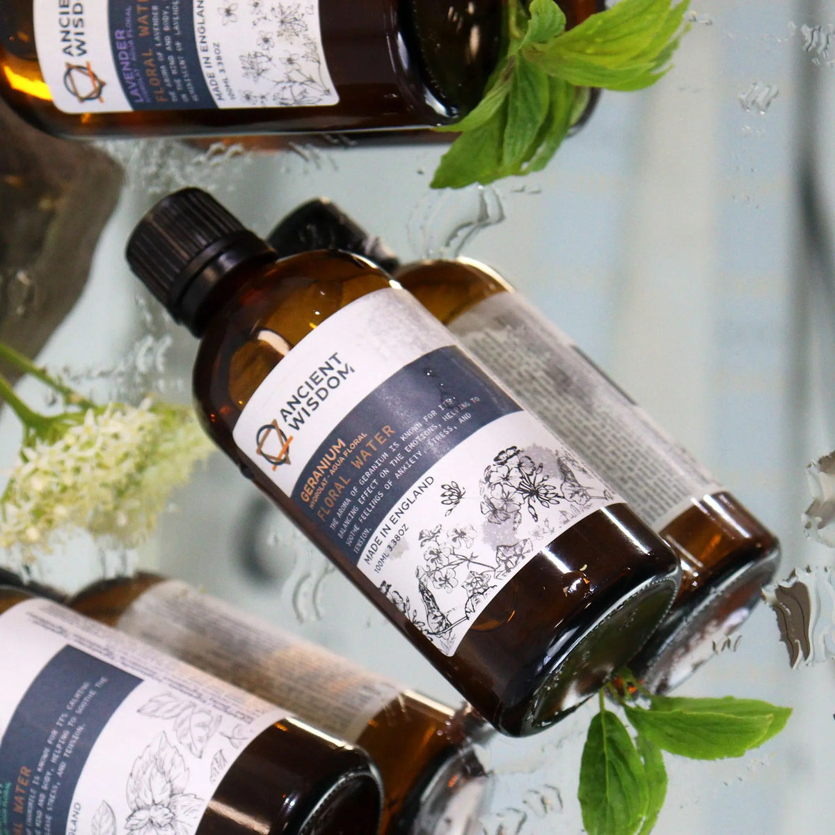 Frasco de vidrio ámbar de 100ml con hidrolato de geranio, sobre superficie reflectante con hojas y flores frescas.