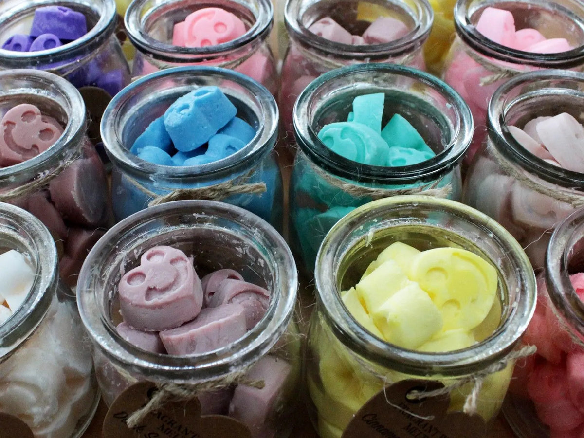 Frascos de vidrio con pequeñas piezas de cera de soja coloridas en forma de caras sonrientes.