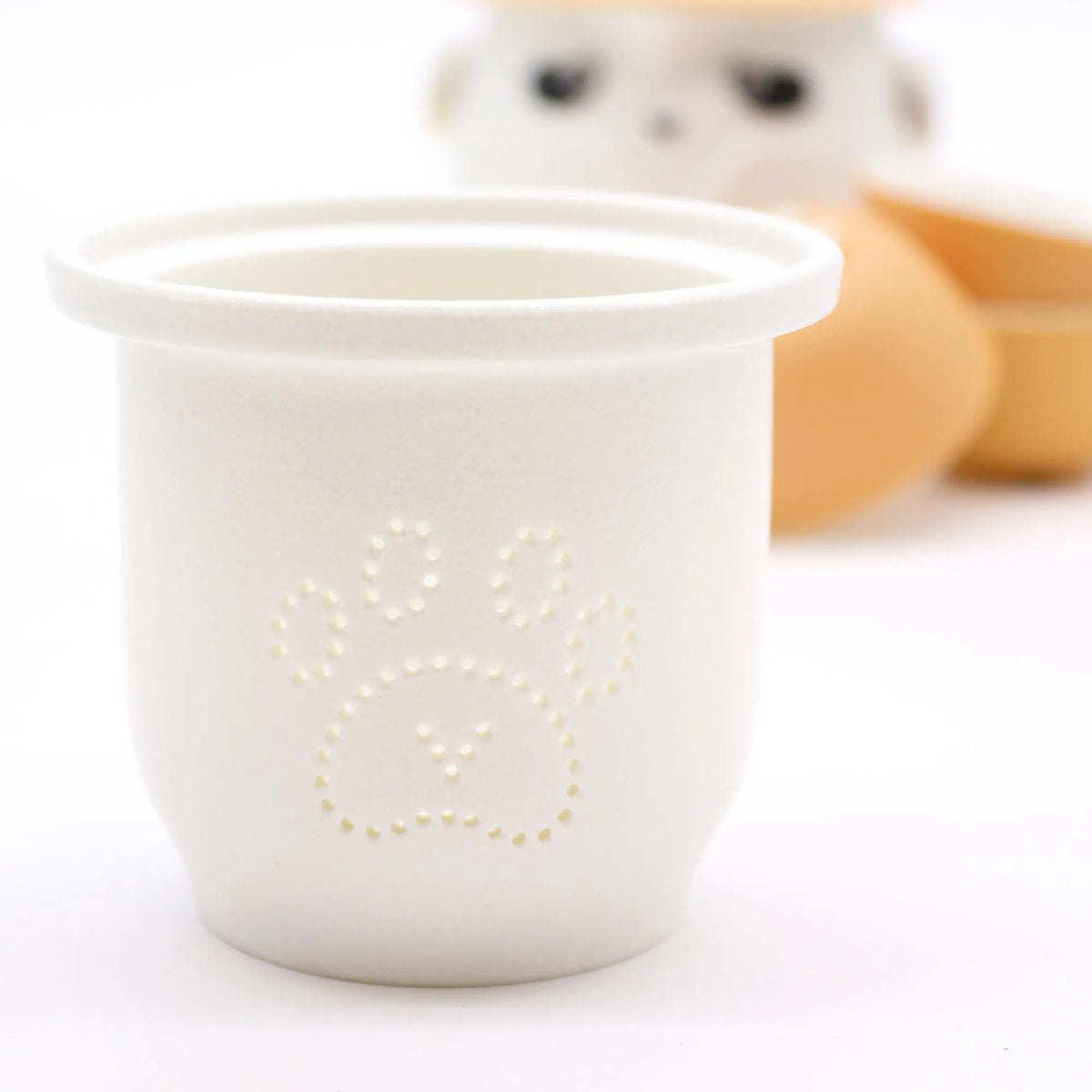 Taza de té blanca de cerámica con huella perforada; al fondo, una tetera de panda y tazas naranjas desenfocadas.
