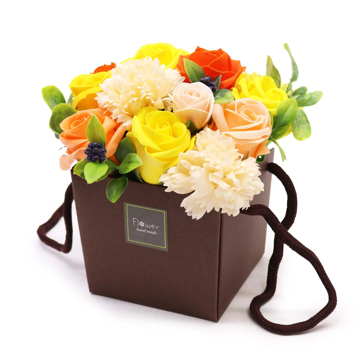 Bouquet de flores de jabón amarillas, naranjas y cremas en caja cuadrada marrón con asas, decoradas con hojas verdes.