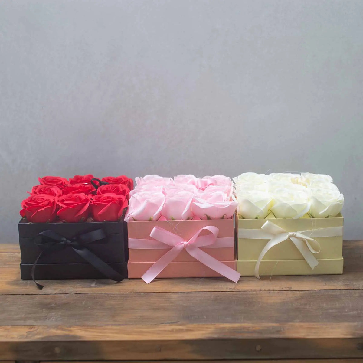 Tres cajas cuadradas de regalo con rosas de jabón rojas, rosas y blancas, decoradas con lazos, sobre una mesa de madera.