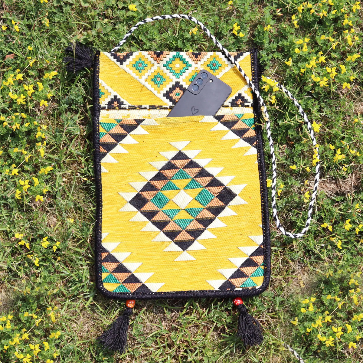 Bolso bandolera de kilim amarillo con patrón geométrico, asa trenzada, borlas negras y un móvil en el bolsillo frontal.