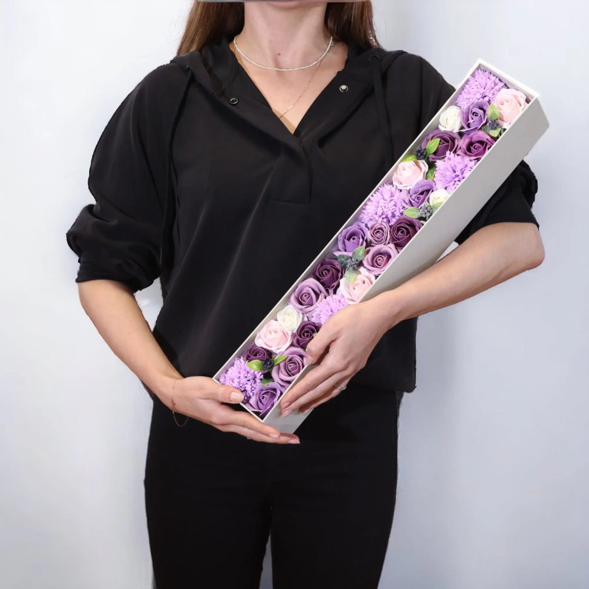 Caja alargada con rosas y claveles de jabón en tonos lavanda, rosa y blanco, sostenida por una persona de negro.