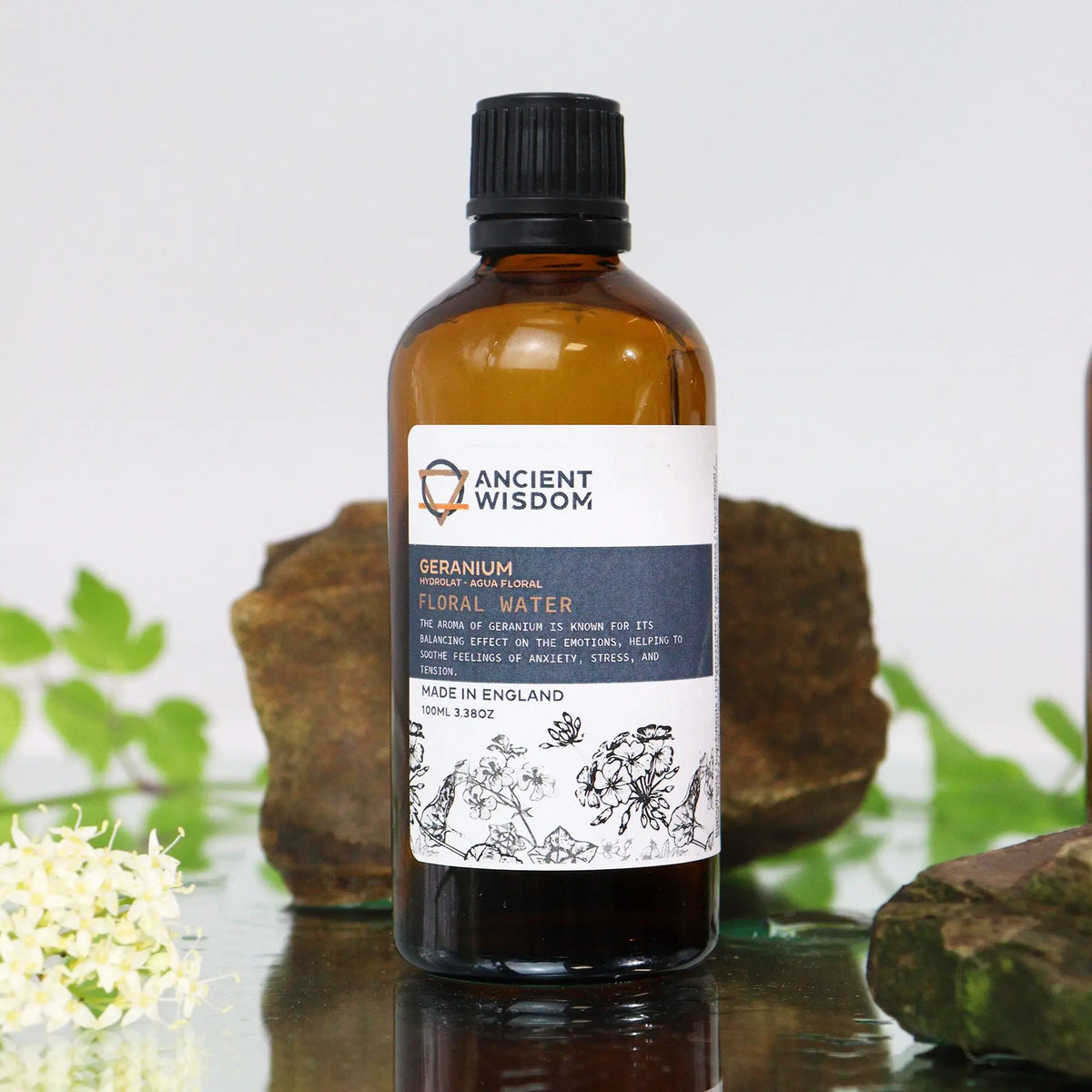 Frasco de vidrio ámbar con agua floral de geranio (100ml), etiqueta ilustrada sobre fondo de rocas y flores naturales.
