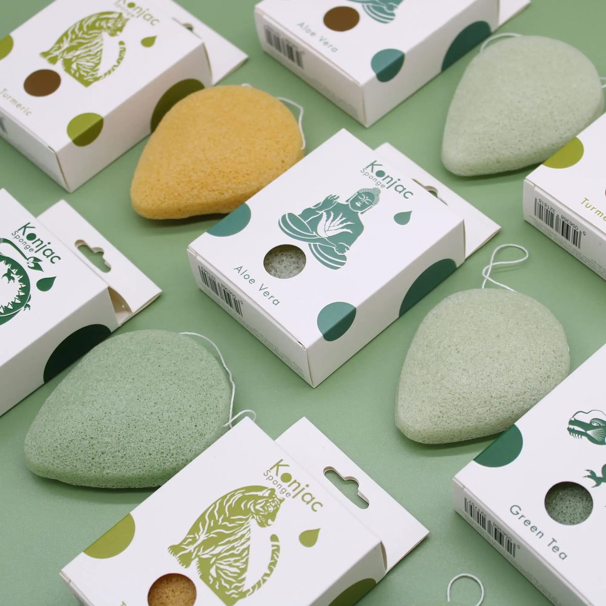 Esponjas faciales Konjac con forma de gota, en tonos verde y amarillo, junto a cajas de cartón sobre fondo verde.