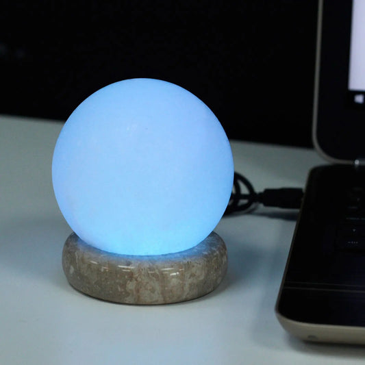 Lámpara de sal blanca esférica con luz LED azul sobre base circular de piedra, conectada a un ordenador vía USB.