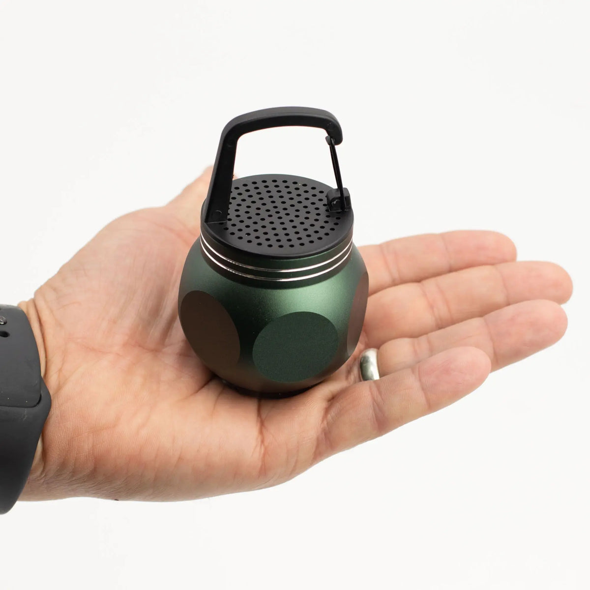 Altavoz Bluetooth mini de metal verde con forma esférica, rejilla negra y gancho superior, sostenido en una mano.