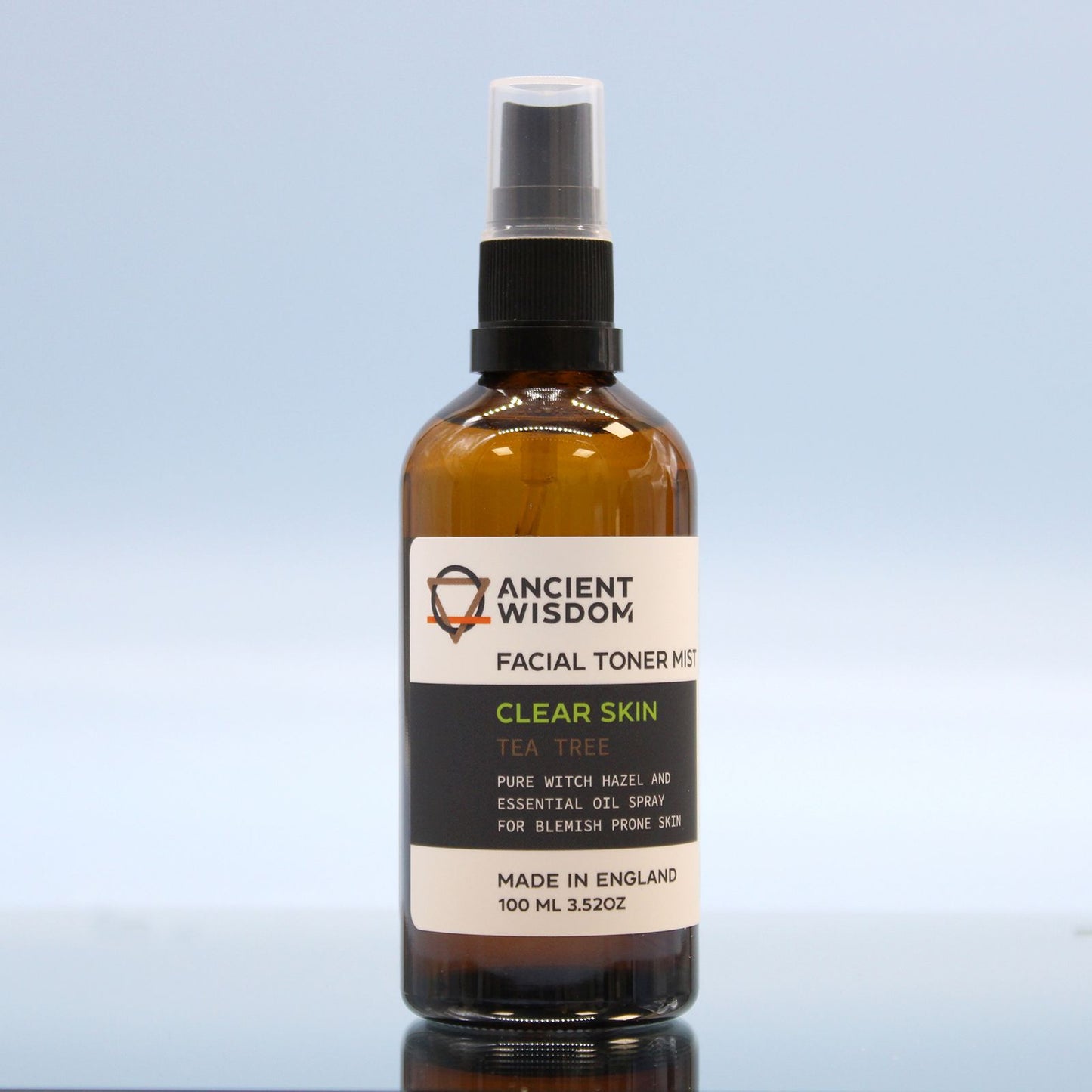 Envase de cristal ámbar con spray, etiqueta blanca y negra de tónico facial de árbol de té, sobre fondo azul claro.