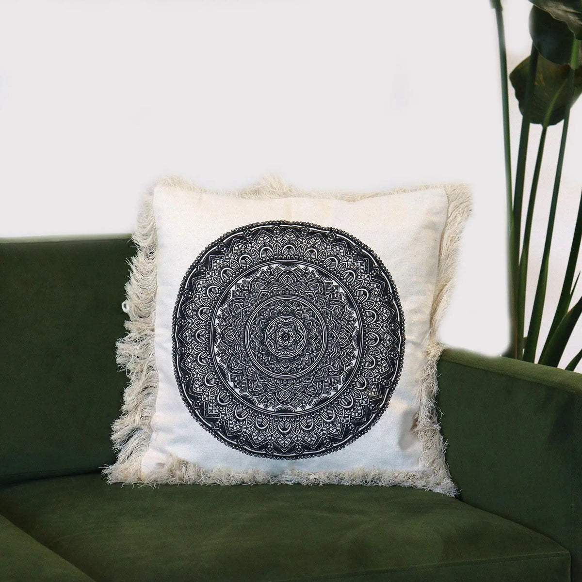 Cojín cuadrado de tela crema con estampado de mandala negro y flecos en los bordes, sobre un sofá verde.
