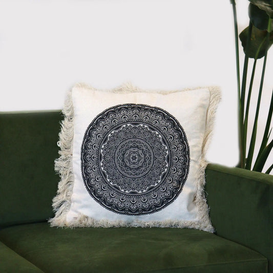 Cojín cuadrado de tela crema con estampado de mandala negro y flecos en los bordes, sobre un sofá verde.
