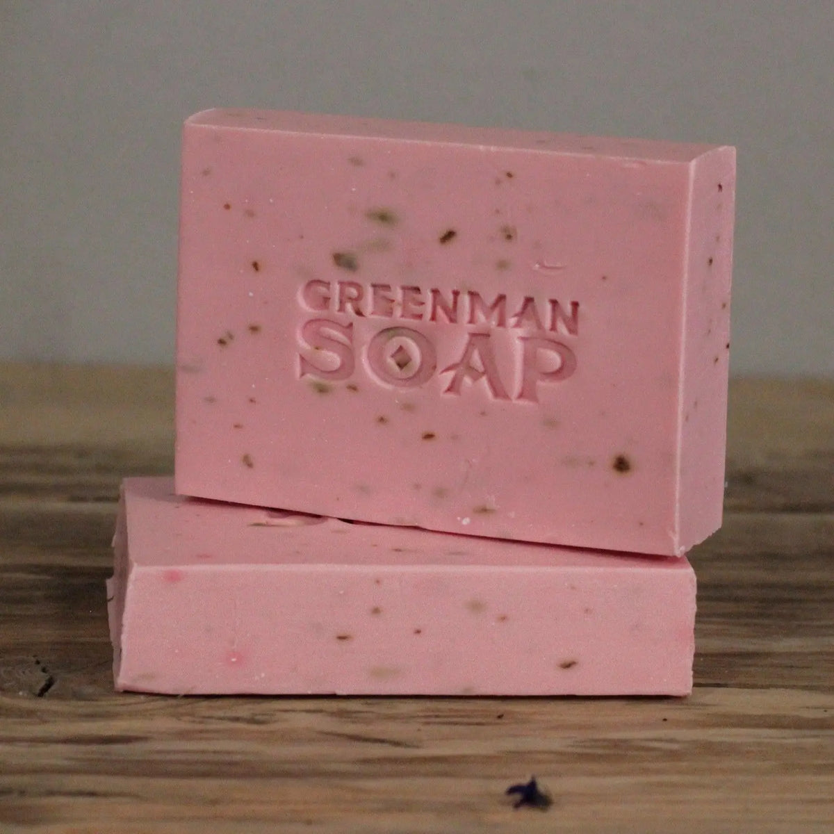 Dos pastillas de jabón rosa con motas, marcadas como "Greenman Soap", apiladas sobre una superficie de madera rústica.