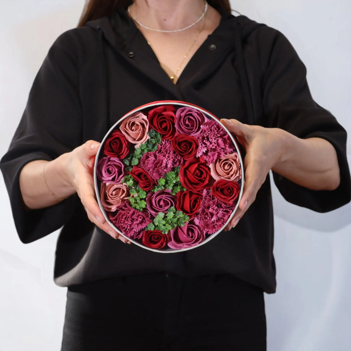 Persona sostiene una caja redonda con rosas de jabón en tonos rojos, rosas y fucsia con detalles de hojas verdes.