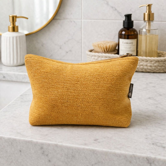 Bolso de maquillaje rectangular en color mostaza, de tela suave, con cremallera dorada y diseño moderno.