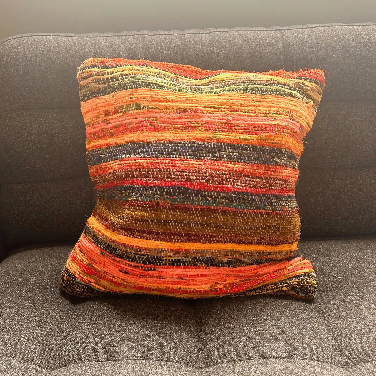 Funda de cojín cuadrada de tejido artesanal en tonos naranja, terracota y rayas oscuras, sobre un sofá gris.