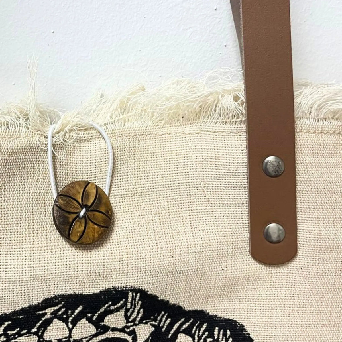 Bolso de tela natural con borde deshilachado, asa de piel marrón con remaches metálicos y botón decorativo de madera.