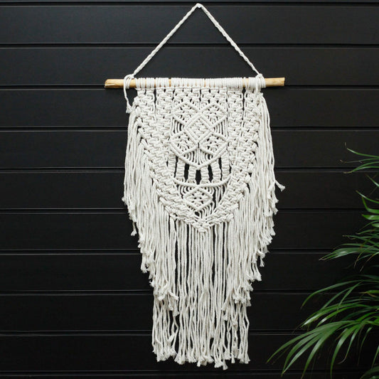 Macramé decorativo blanco con flecos en forma de diamante, colgado en una barra de madera. Ideal para interiores.